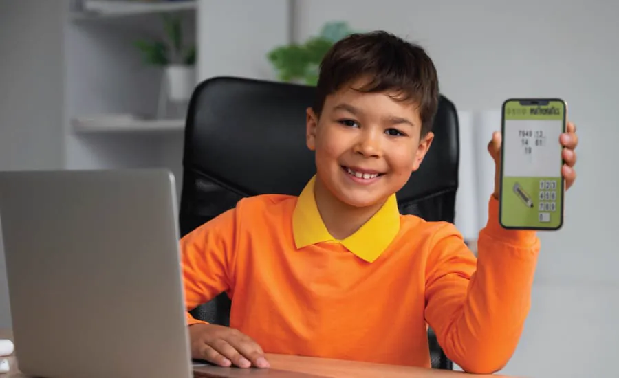 A boy holds up a phone showing the app he made, with his laptop beside him