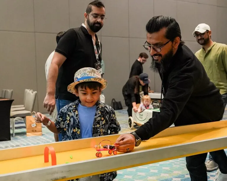 A young boy smiles as he races a small robot car down a track, guided by an adult