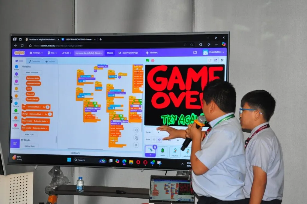 Two students pitch the game they built at Code Battle 2024, with their code on the big screen