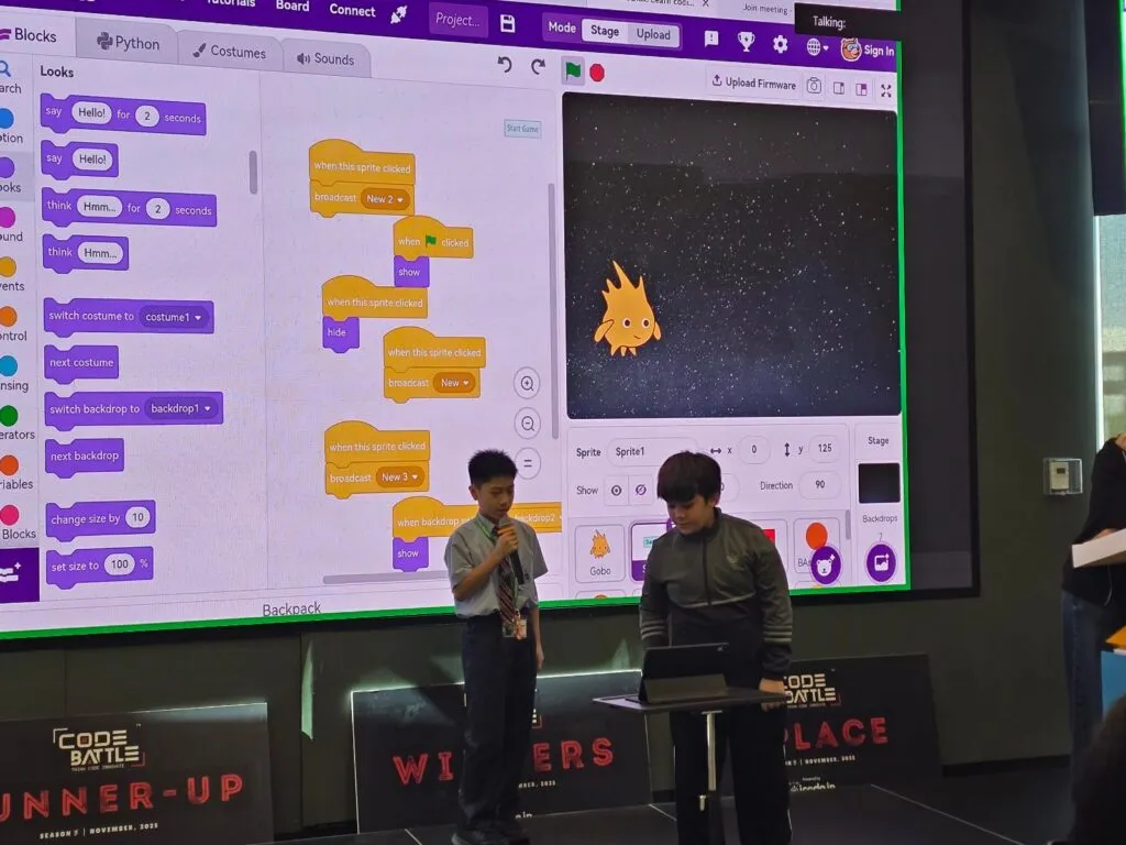 Two students presenting a block-coding project on a big screen at Code Battle 2025